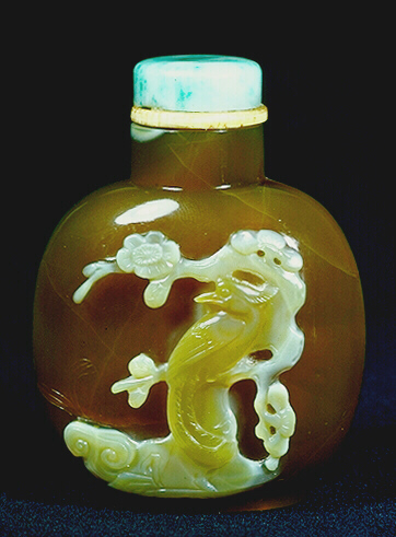 Chalcedony three colour snuff bottle; dark mottled brown with an opaque white area topped by a caramel inclusion, one side carved with a duck relaxing on a large lotus leaf, the other with a phoenix against a flowering prunus, with ruyi.